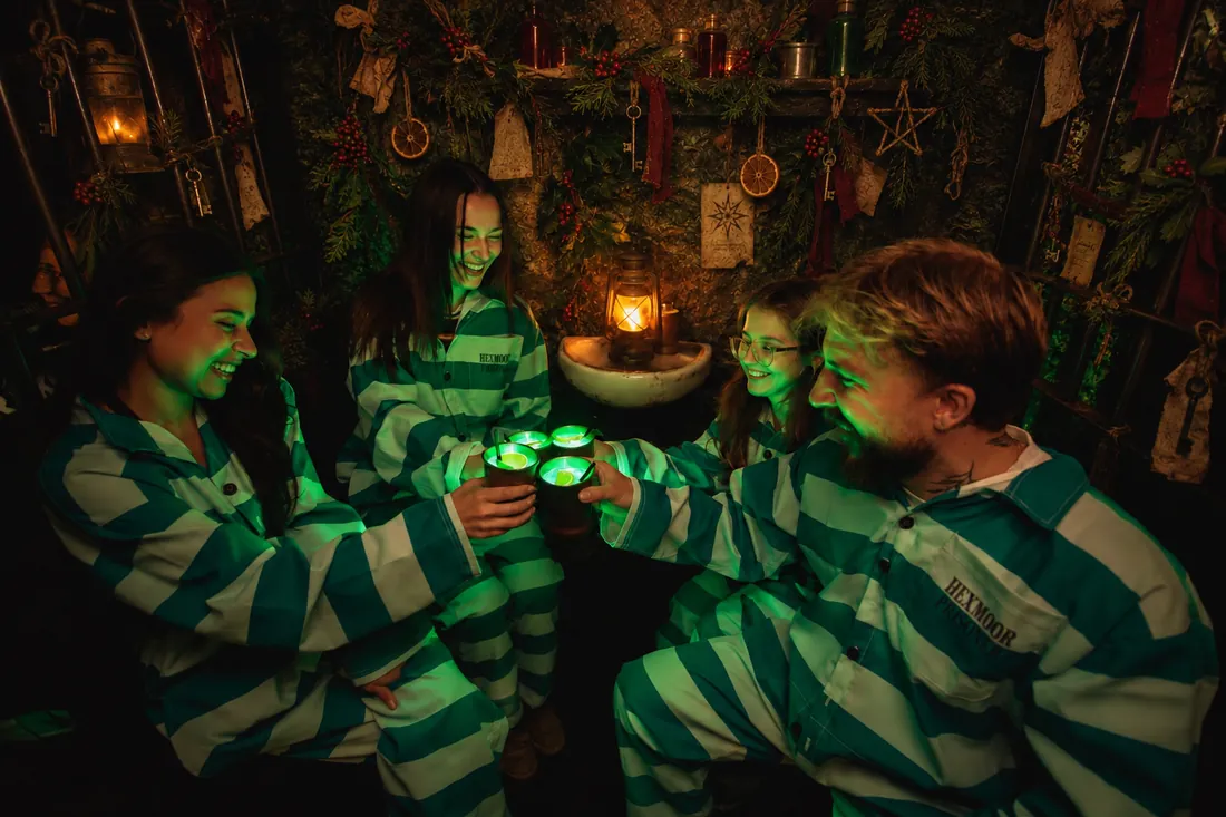Four guests in green striped jumpsuits toasting with glowing cocktails in a decorated cell