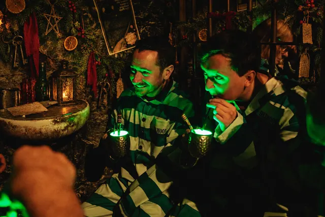 Two guests in jumpsuits drinking glowing green cocktails in a candlelit cell